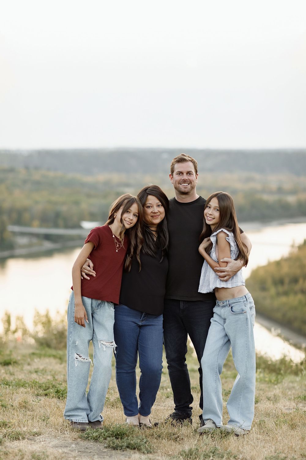 Family of four smiling together on a hilltop with a scenic river valley backdrop on a warm day.
