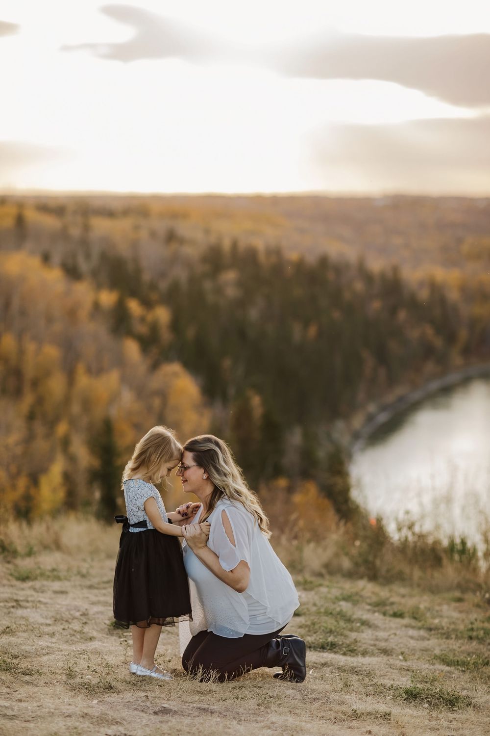 Pregnant mother kneeling on a hilltop sharing a tender moment with her young daughter amid autumn foliage.