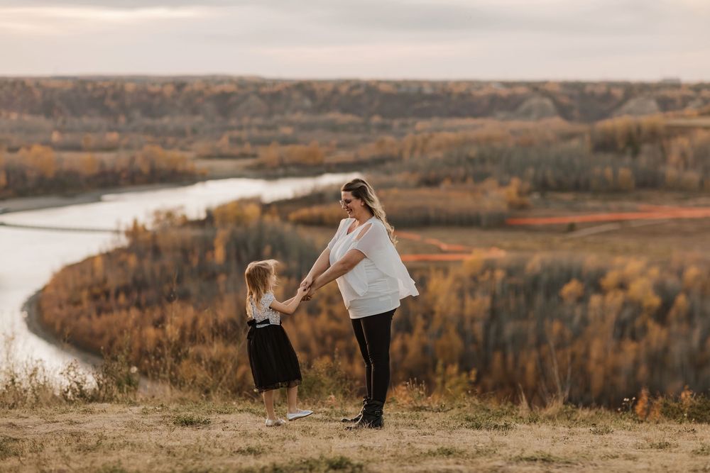 A mother and young daughter hold hands on a hilltop overlooking a winding river and autumn foliage landscape.