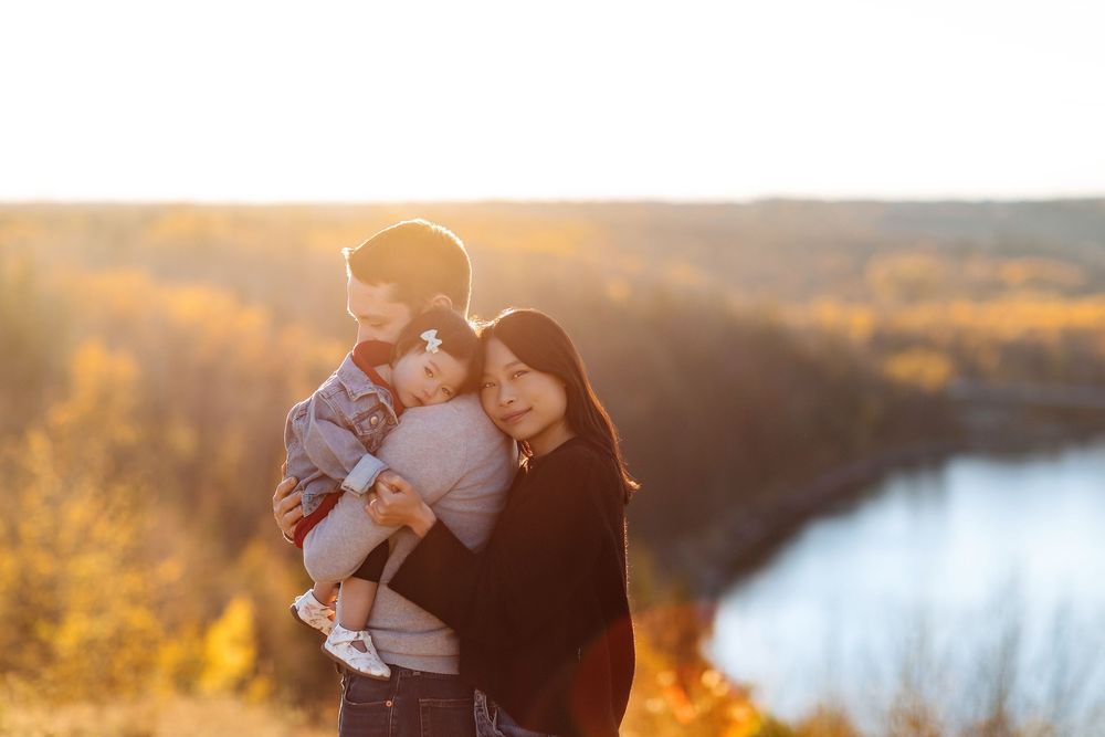 A young family with a baby enjoys a golden sunset overlooking a river and autumn foliage.