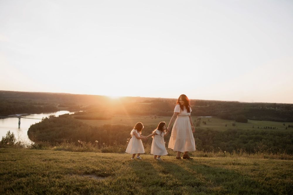 A mother and two daughters in white dresses walk through a sunlit meadow with a river valley at golden hour.