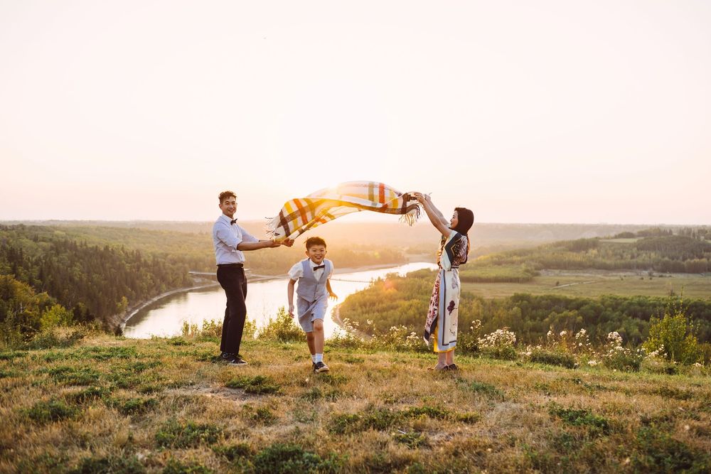 Family of three playing with a colorful blanket on a hilltop at sunset with a river valley in the background.