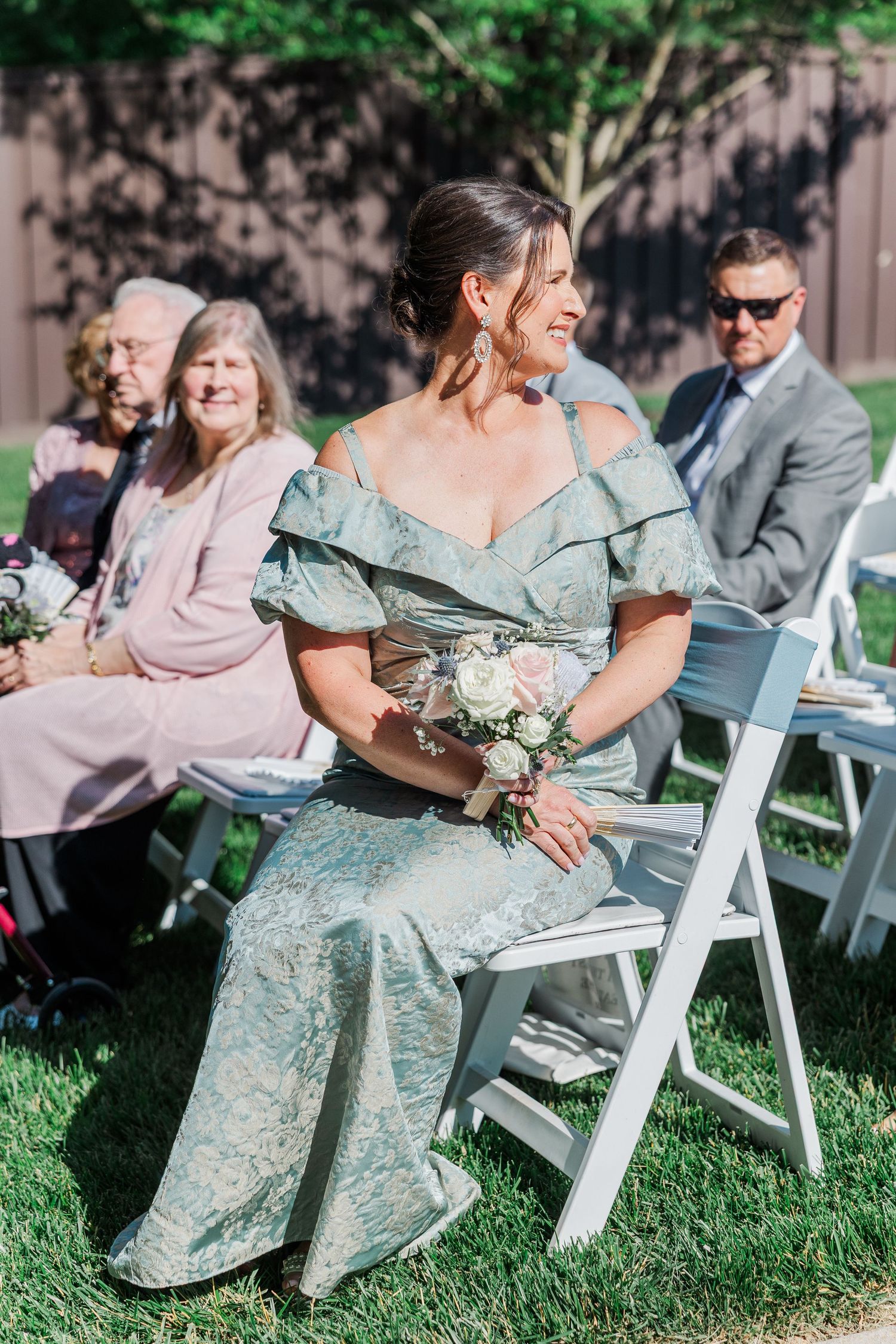 Bridesmaid in sage green off-shoulder gown holds bouquet at outdoor wedding ceremony with guests seated on white chairs.