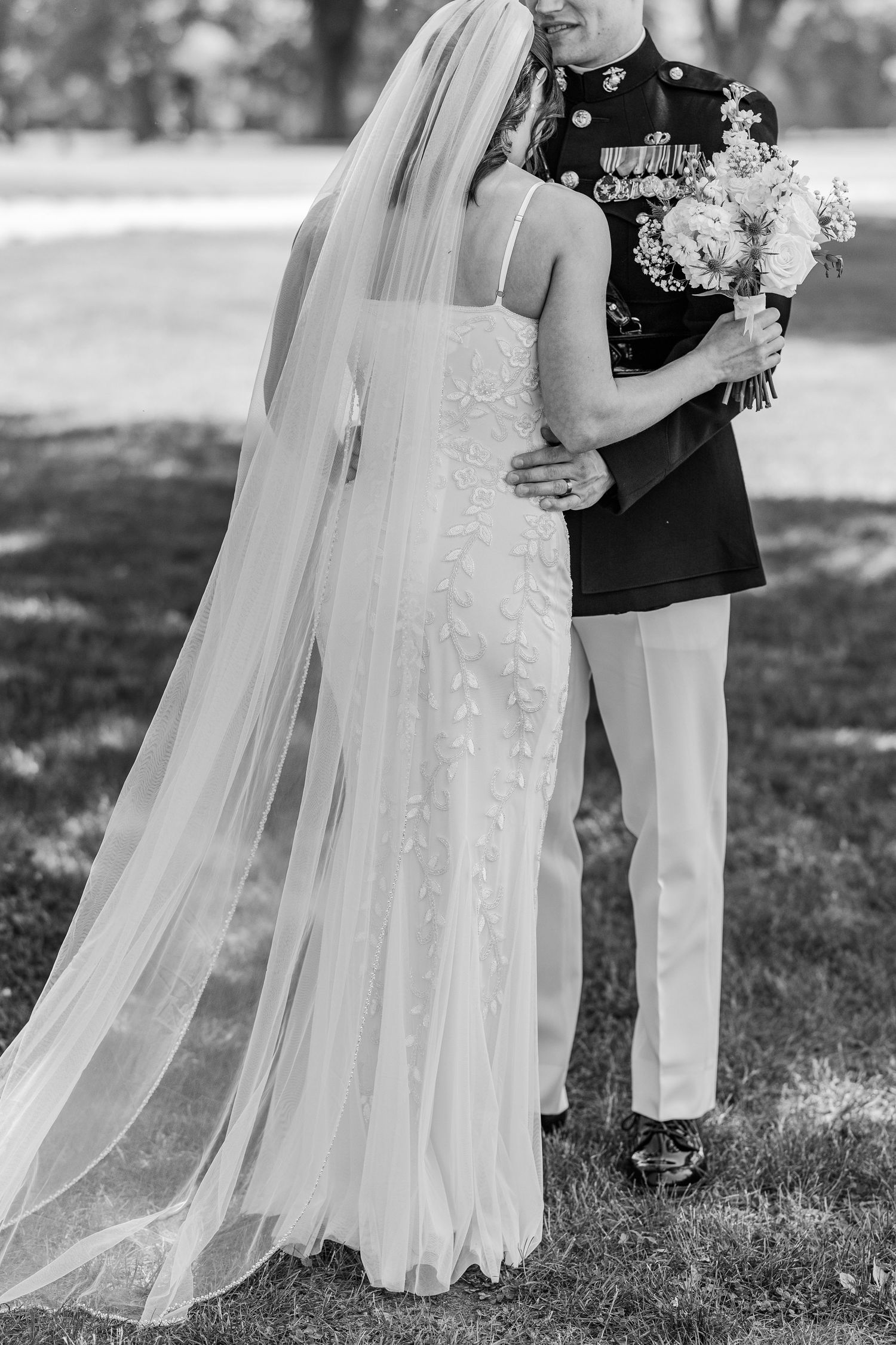 Black and white photo of a bride in a lace gown and veil with a groom in military dress uniform holding a bouquet.