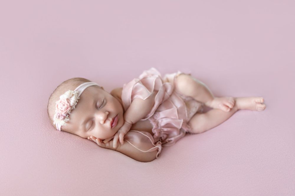Sleeping newborn baby girl in pink outfit and floral headband posed on pink backdrop.