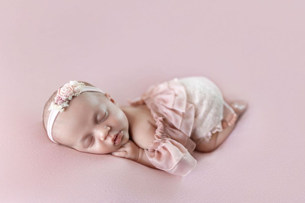 Newborn baby girl sleeping peacefully in pink ruffled outfit and floral headband on soft pink backdrop.