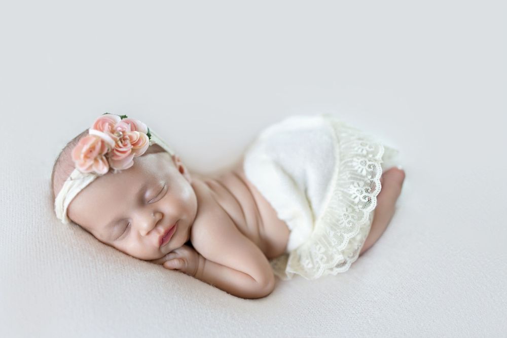 Sleeping newborn baby girl in white lace outfit and pink floral headband on soft white background.