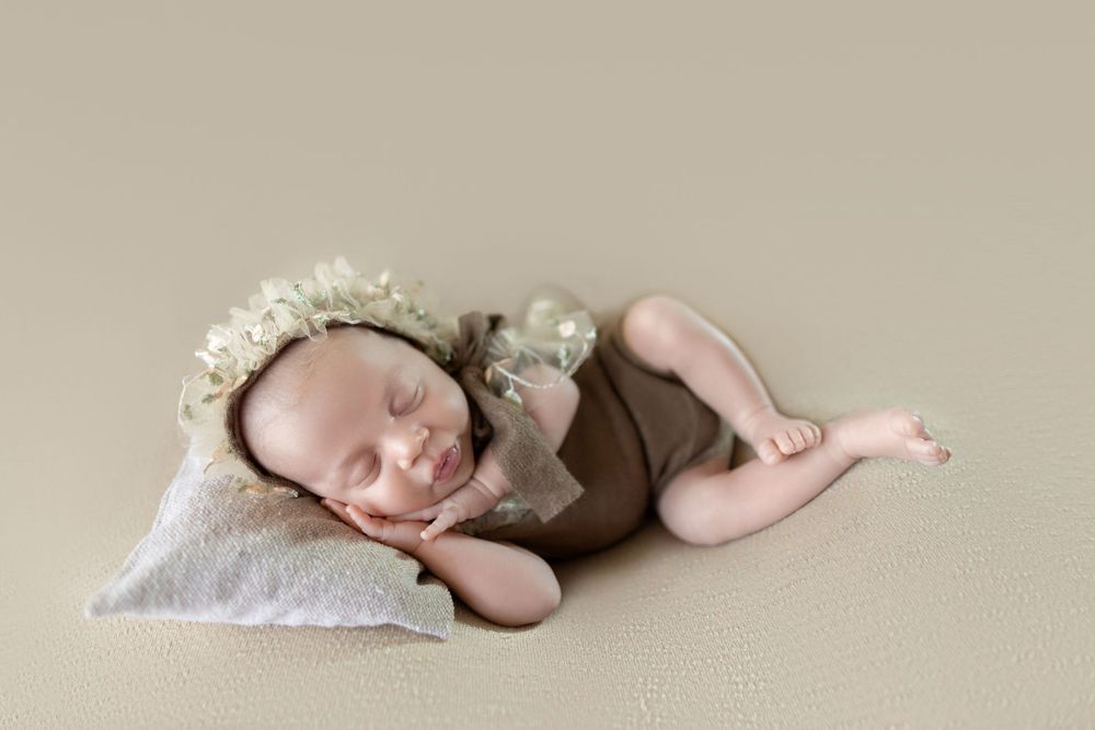Sleeping newborn baby wearing a floral bonnet and brown wrap, posed on a cream pillow against a beige backdrop.