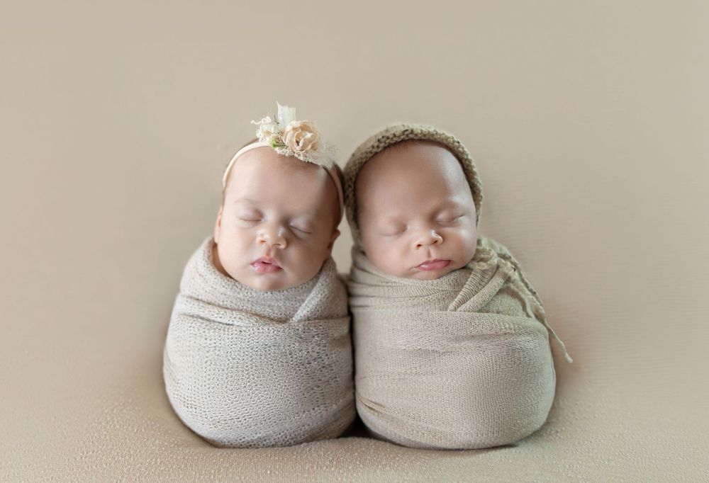 Two swaddled newborn twins sleeping side by side, one with a floral headband, one in a green bonnet.