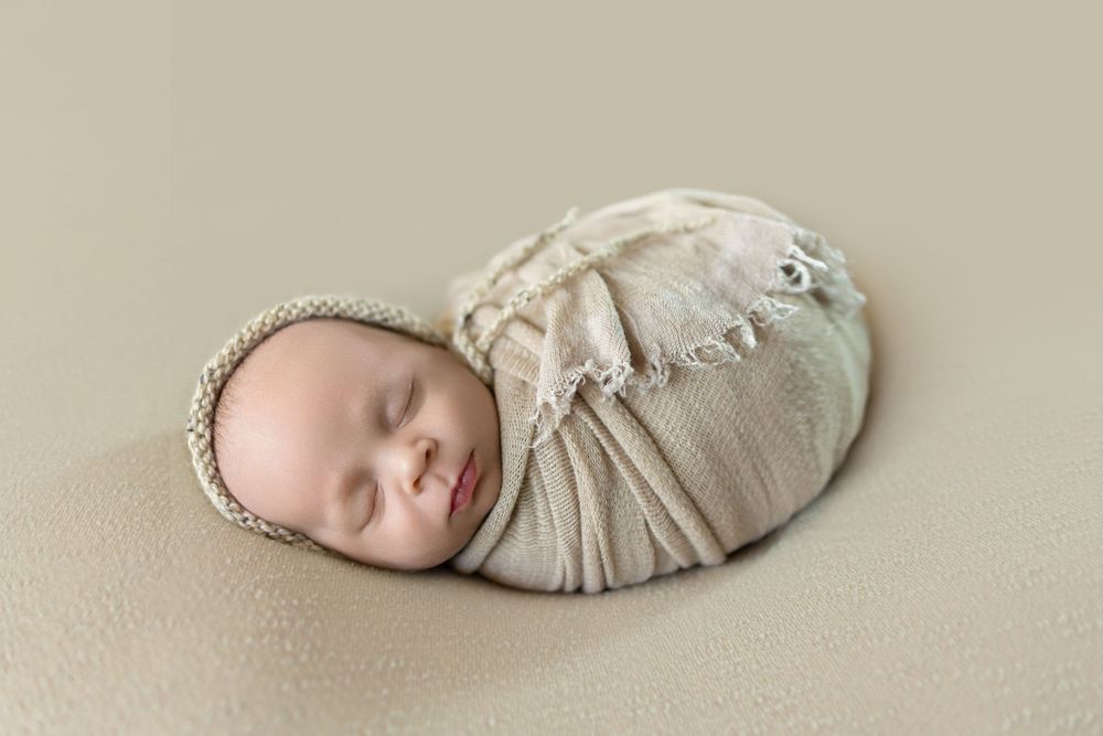 Sleeping newborn baby wrapped in cream knit swaddle blanket wearing a bonnet on a beige background.