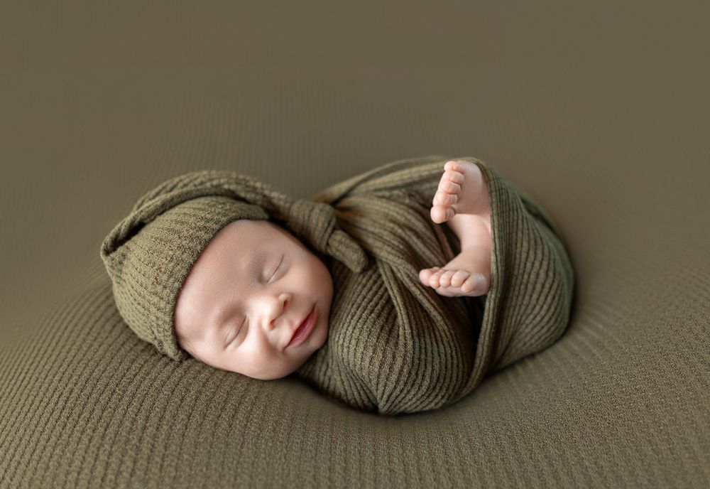 Sleeping newborn baby wrapped in olive green knit blanket and matching hat on green background.