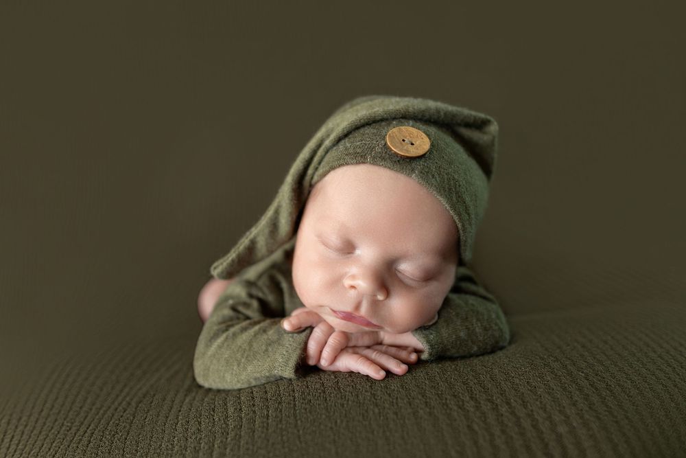 Newborn baby sleeping peacefully in green knit outfit and elf hat with button on olive green background.