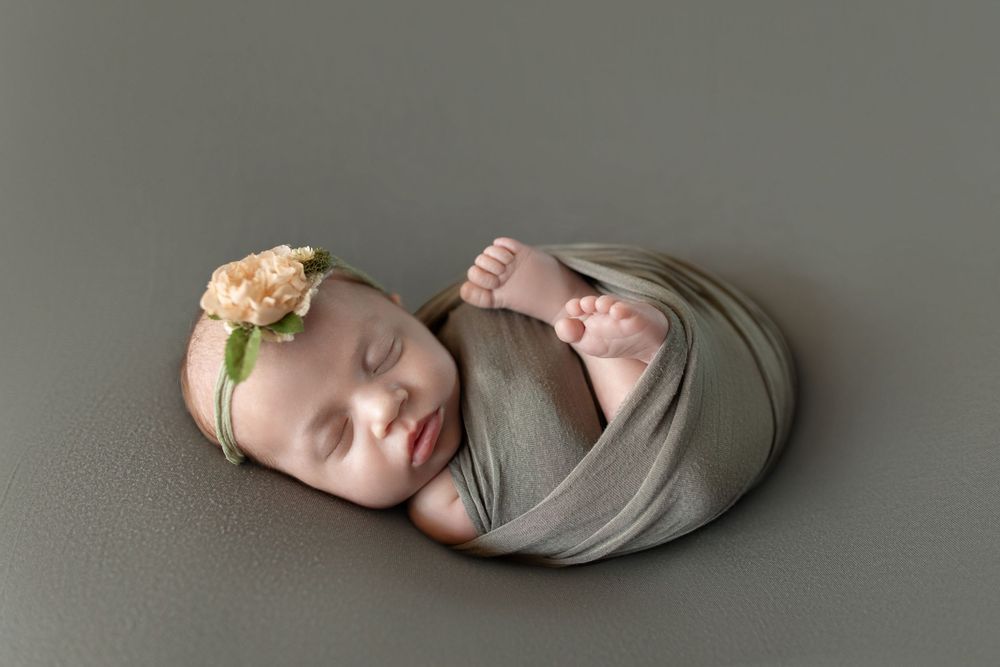 Newborn baby girl swaddled in sage green wrap with floral headband, sleeping peacefully in professional studio photo.