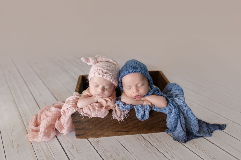 Two sleeping newborn twins in pink and blue knit hats rest together in a wooden box on a white floor.