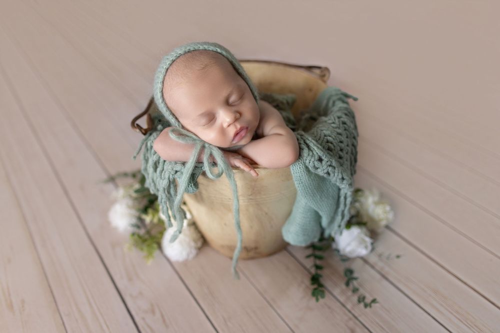 Newborn baby sleeping peacefully in a wooden bowl with teal knit bonnet and blanket, surrounded by white flowers.