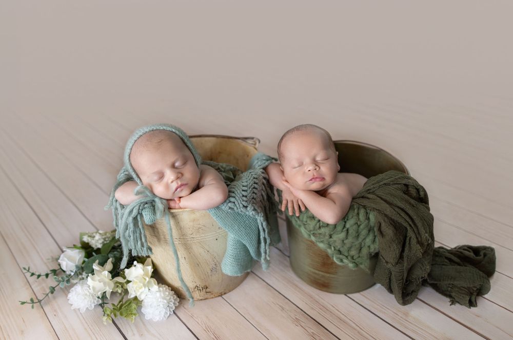Two sleeping newborn twins rest in rustic wooden buckets with soft green wraps and a floral arrangement nearby.