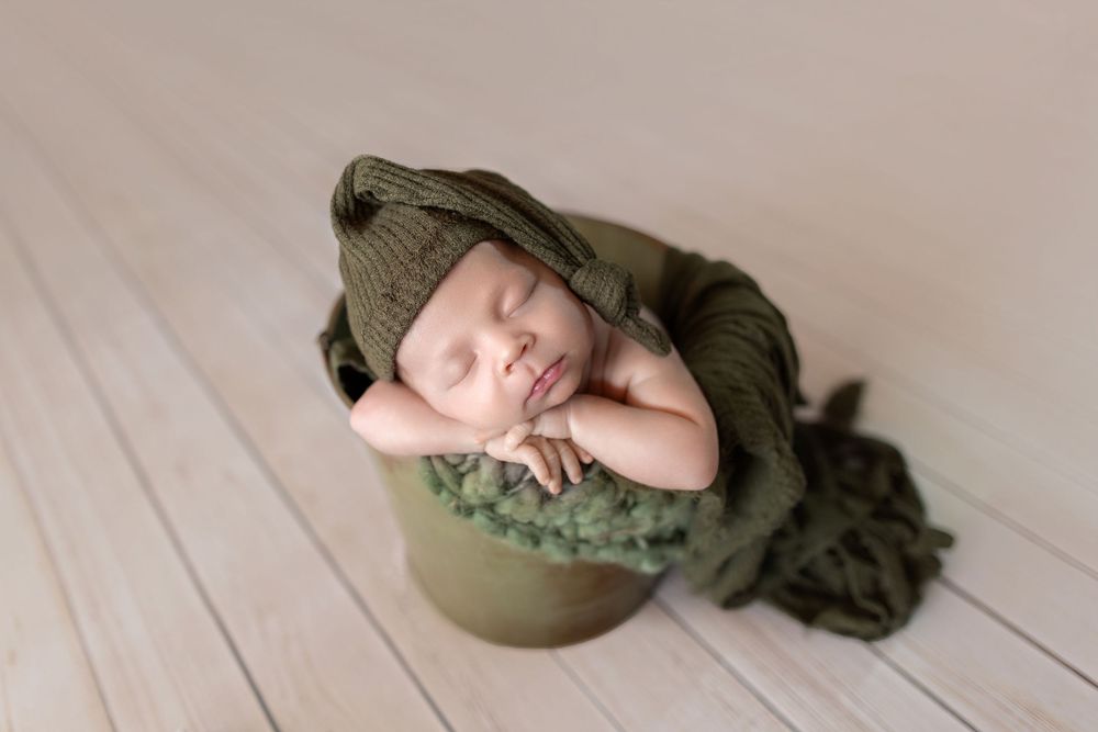 Sleeping newborn baby wrapped in green knit blanket and hat, posed in a green bucket on light wood floor.