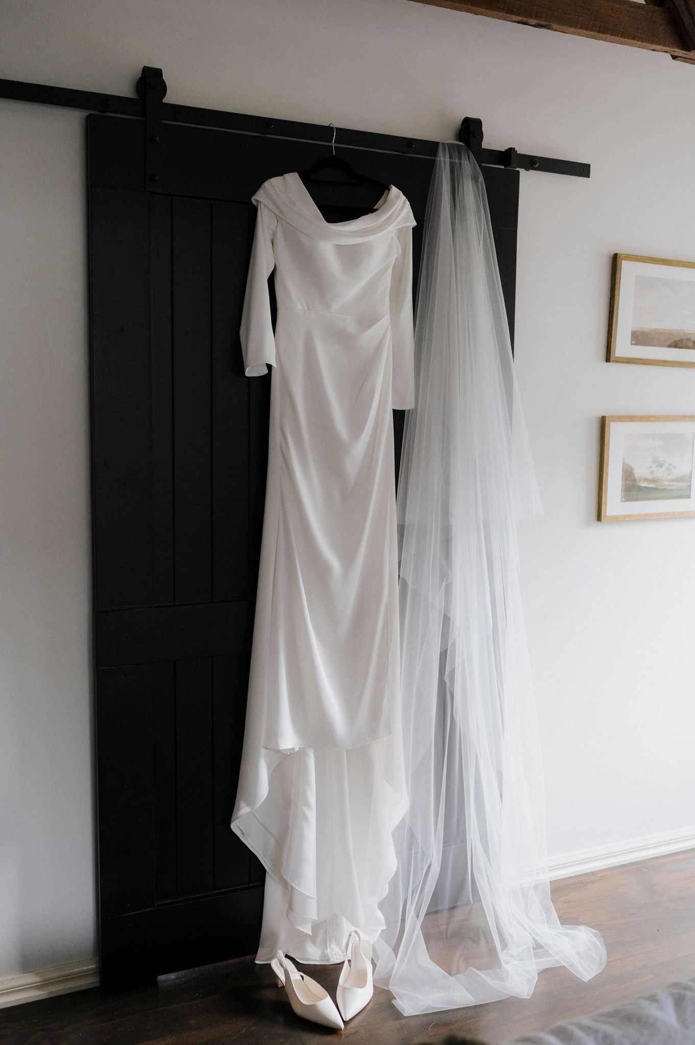 Elegant white long-sleeve wedding dress with flowing veil hanging on a black barn door, with bridal heels below.