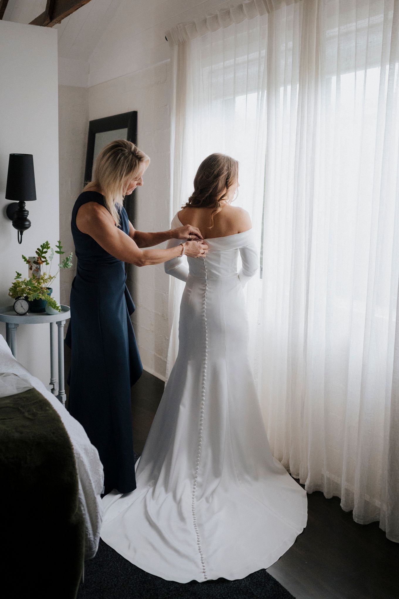 Bride in white off-shoulder gown being helped with buttons by woman in navy dress near sheer curtained window.