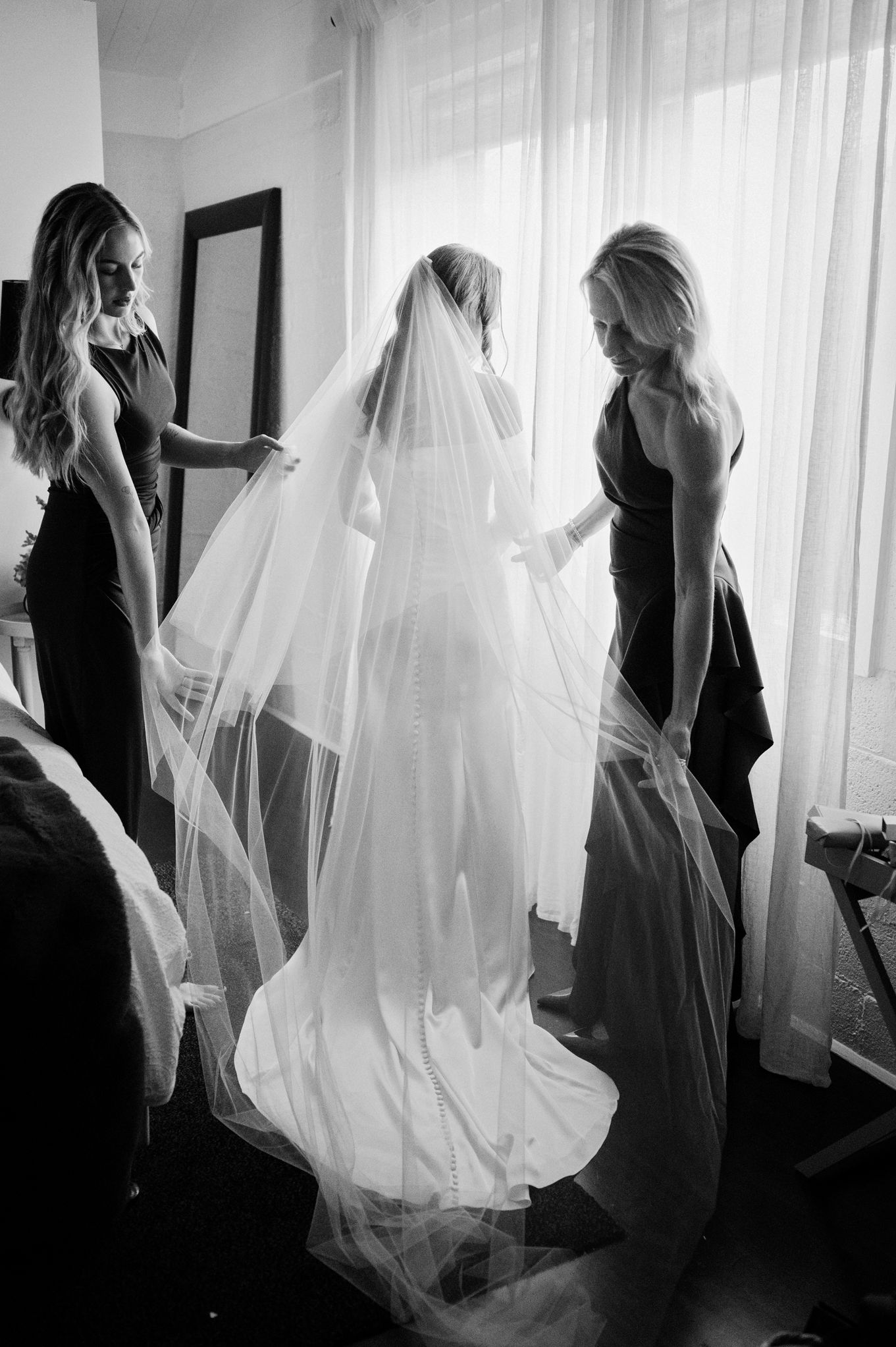 Black and white photo of a bride in a flowing veil and gown, assisted by two bridesmaids in dark dresses near a window.
