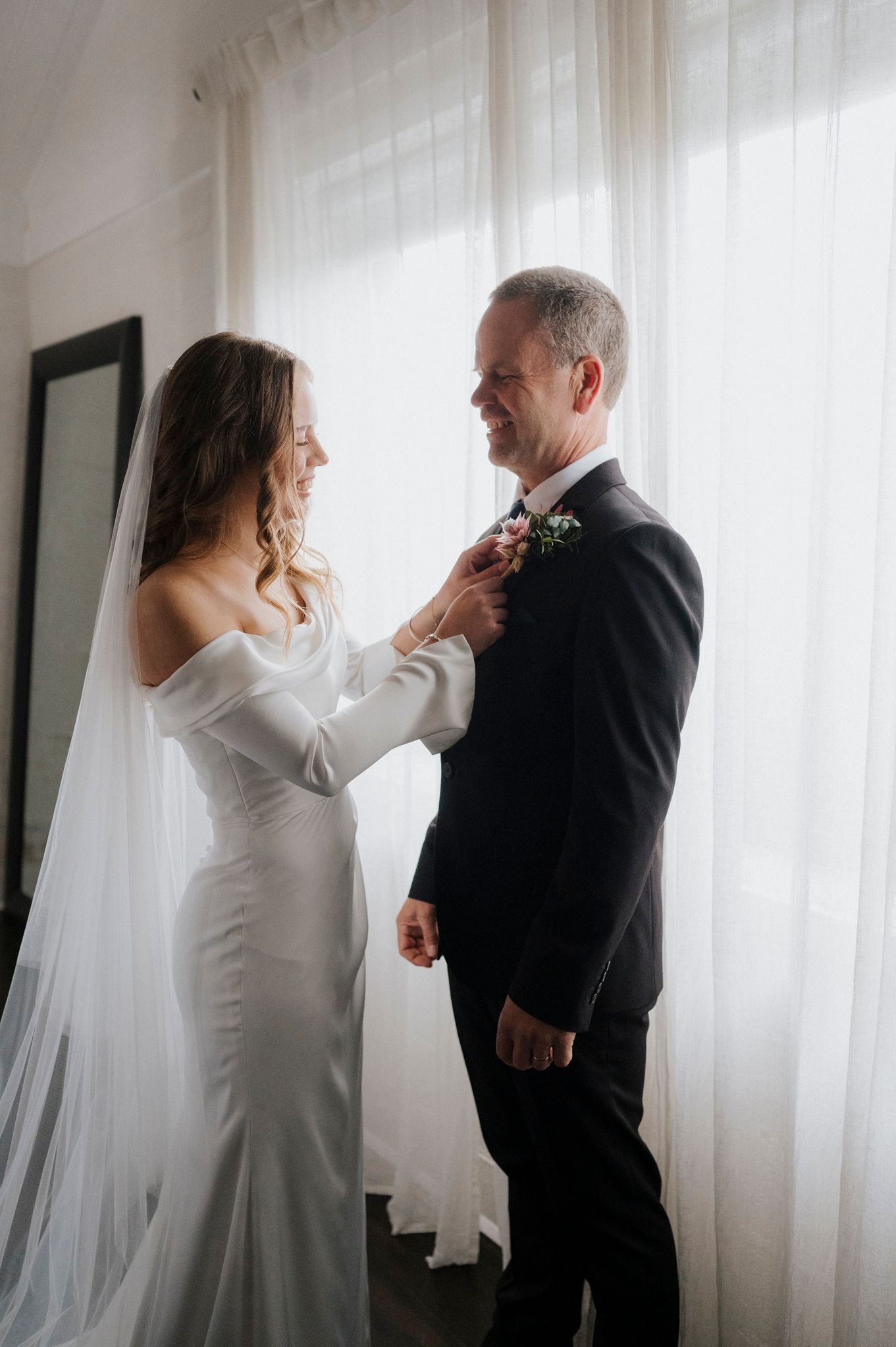 Bride in white off-shoulder gown and veil pins boutonniere on groom in black suit near sheer curtains.