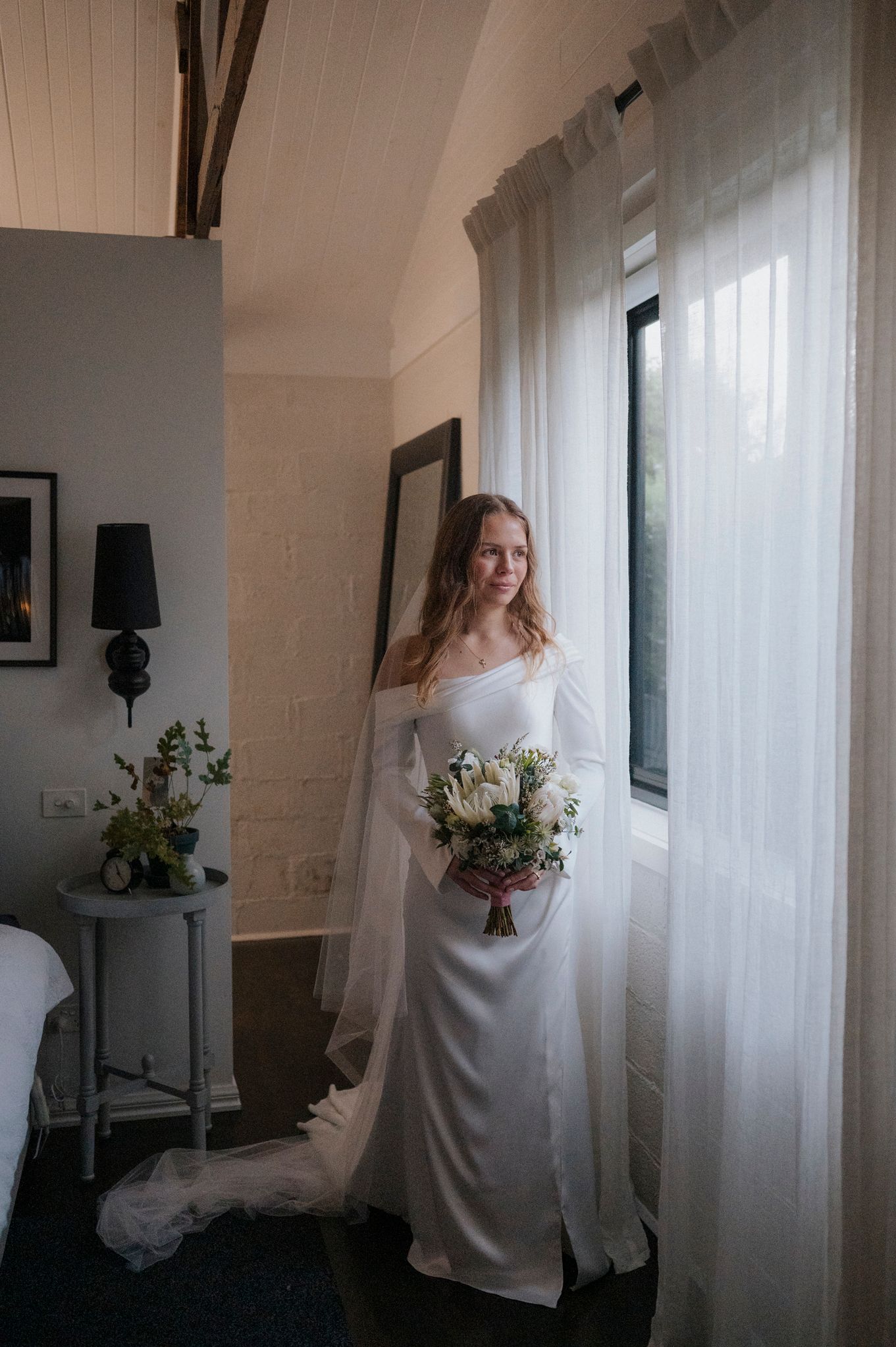 Bride in elegant off-shoulder white gown holding floral bouquet standing by sheer curtained window in softly lit room.