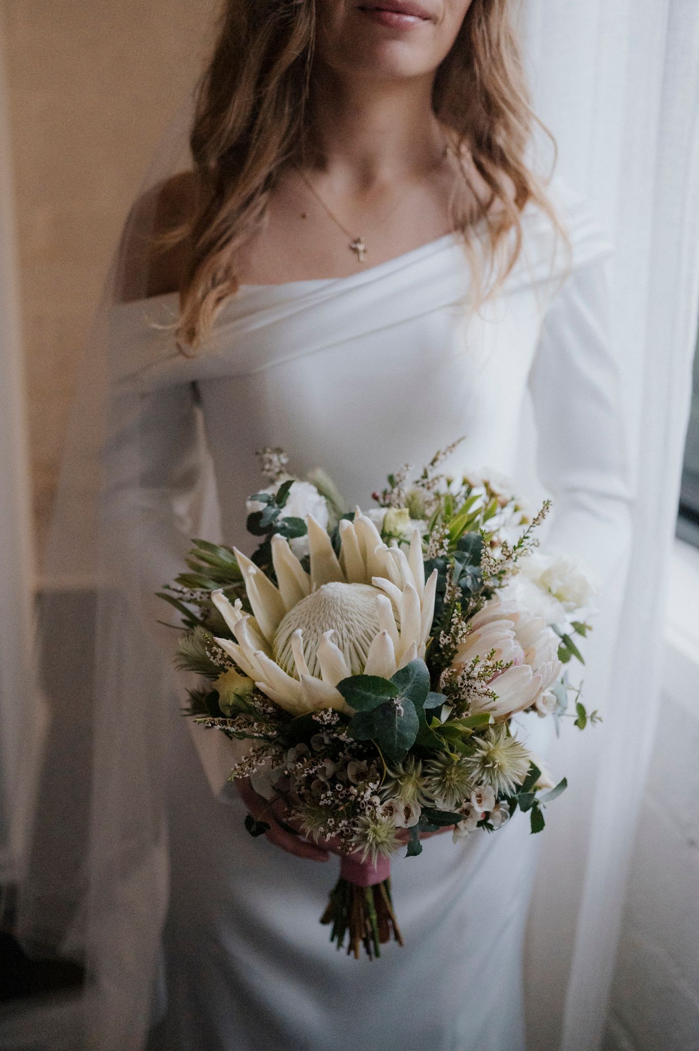 Bride in off-shoulder white gown holding elegant bouquet with king protea, white roses, and greenery.