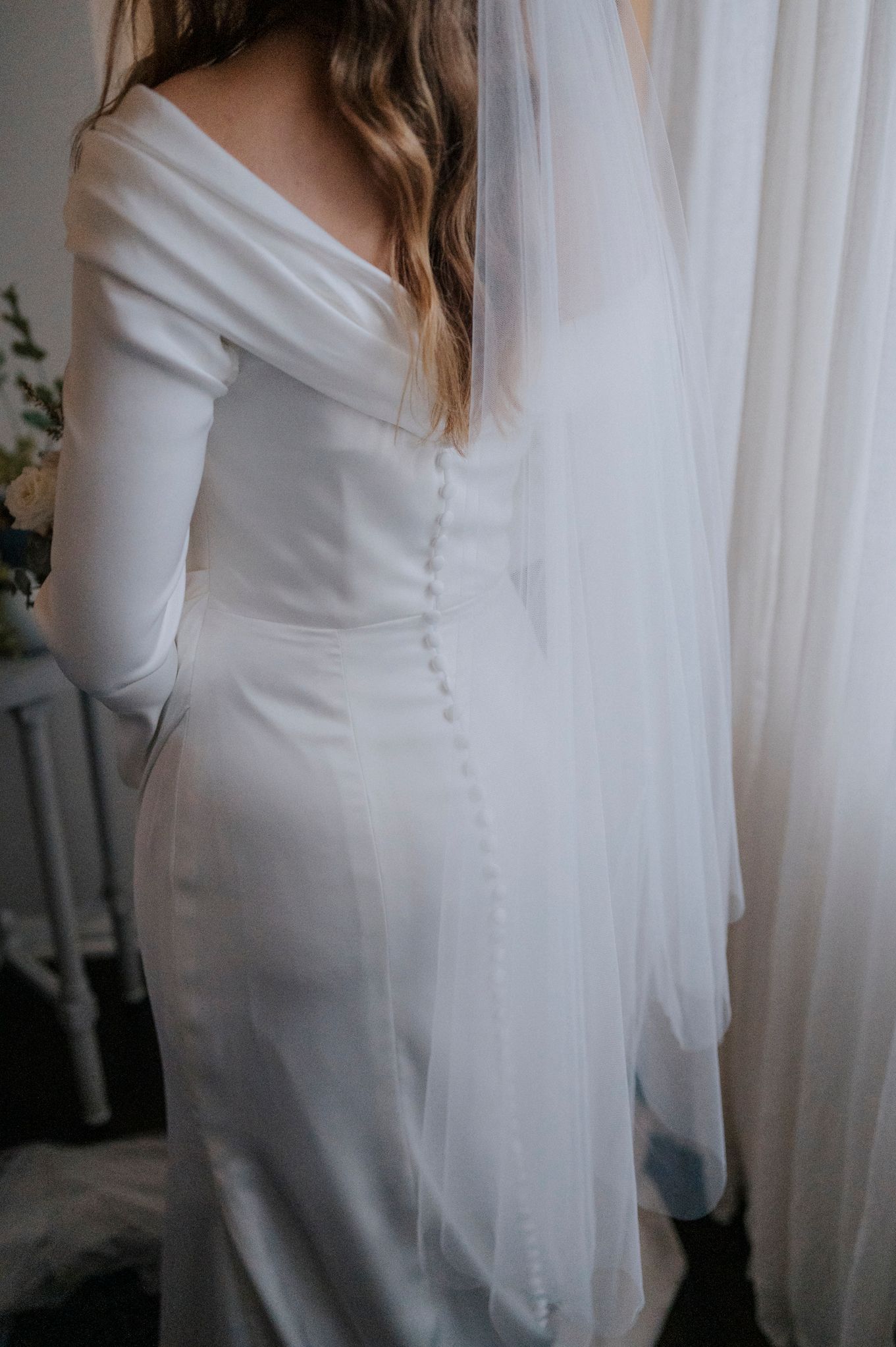Back view of bride in fitted white wedding gown with button detail and flowing veil.
