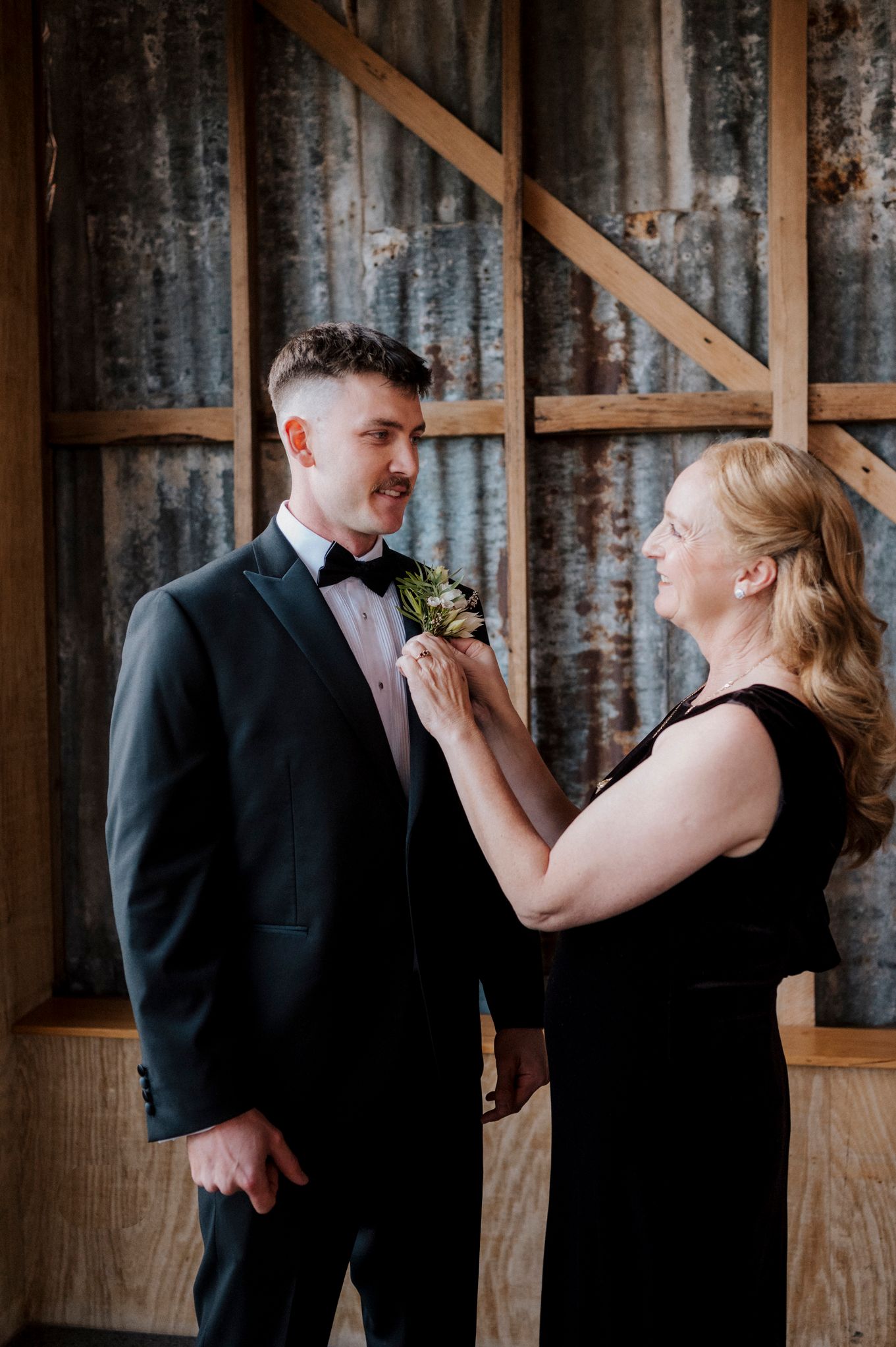 A woman in black dress pins a boutonniere on a groom in a tuxedo inside a rustic barn venue.