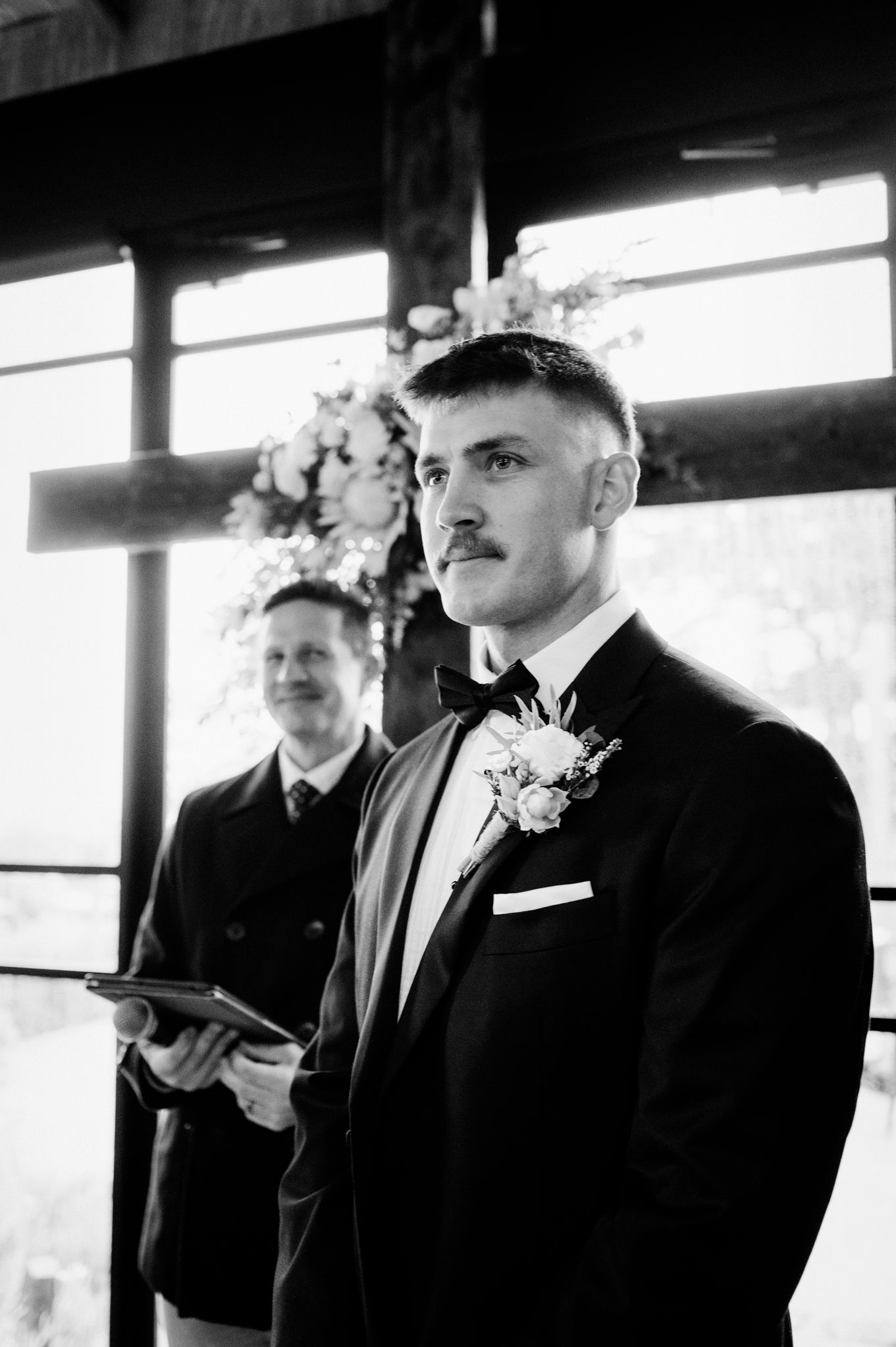 Groom in black tuxedo with boutonniere stands at wedding altar as officiant holds tablet in background.