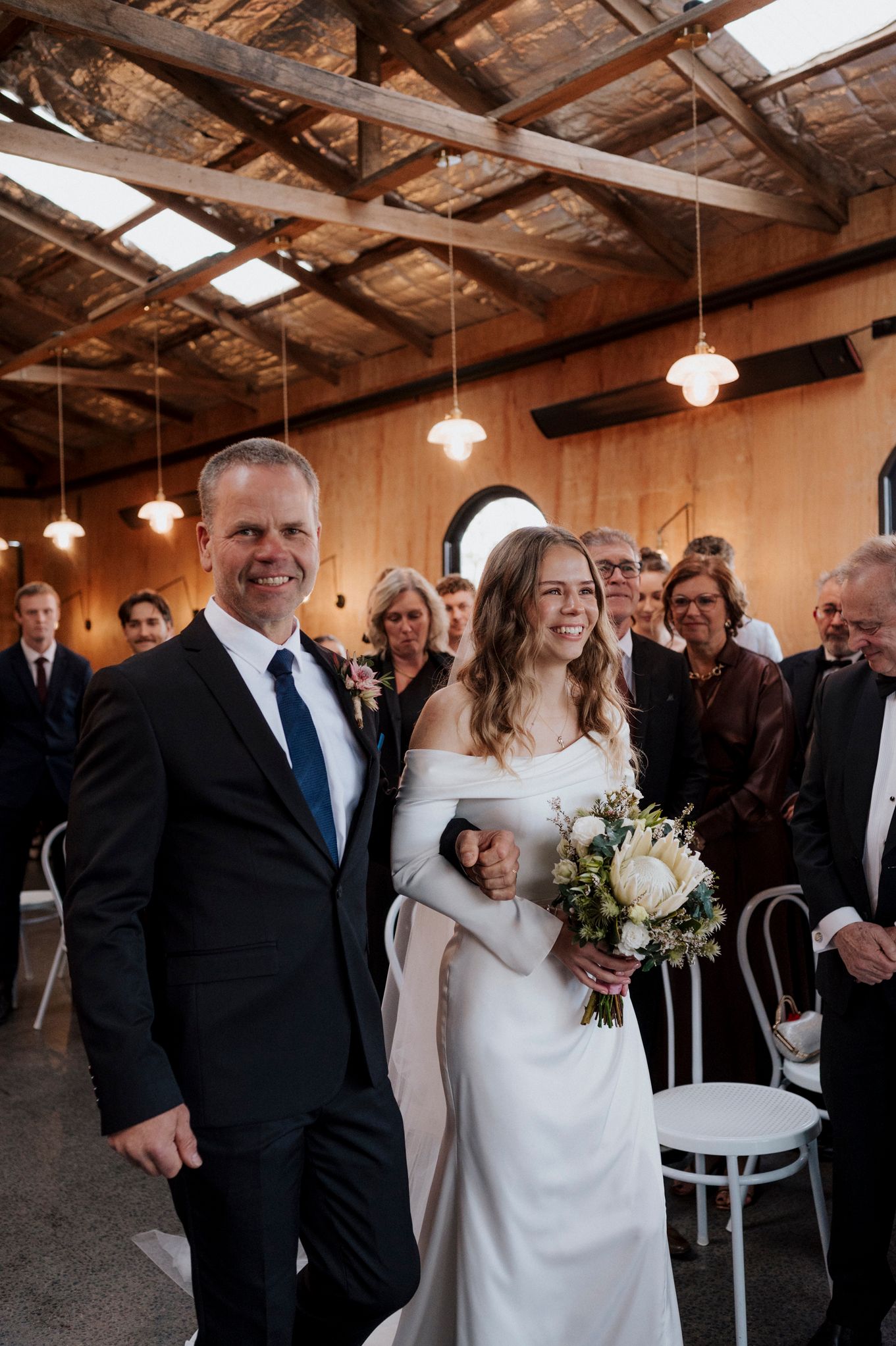 Bride and groom walk down the aisle smiling, bride holds white floral bouquet in elegant off-shoulder gown at indoor venue.