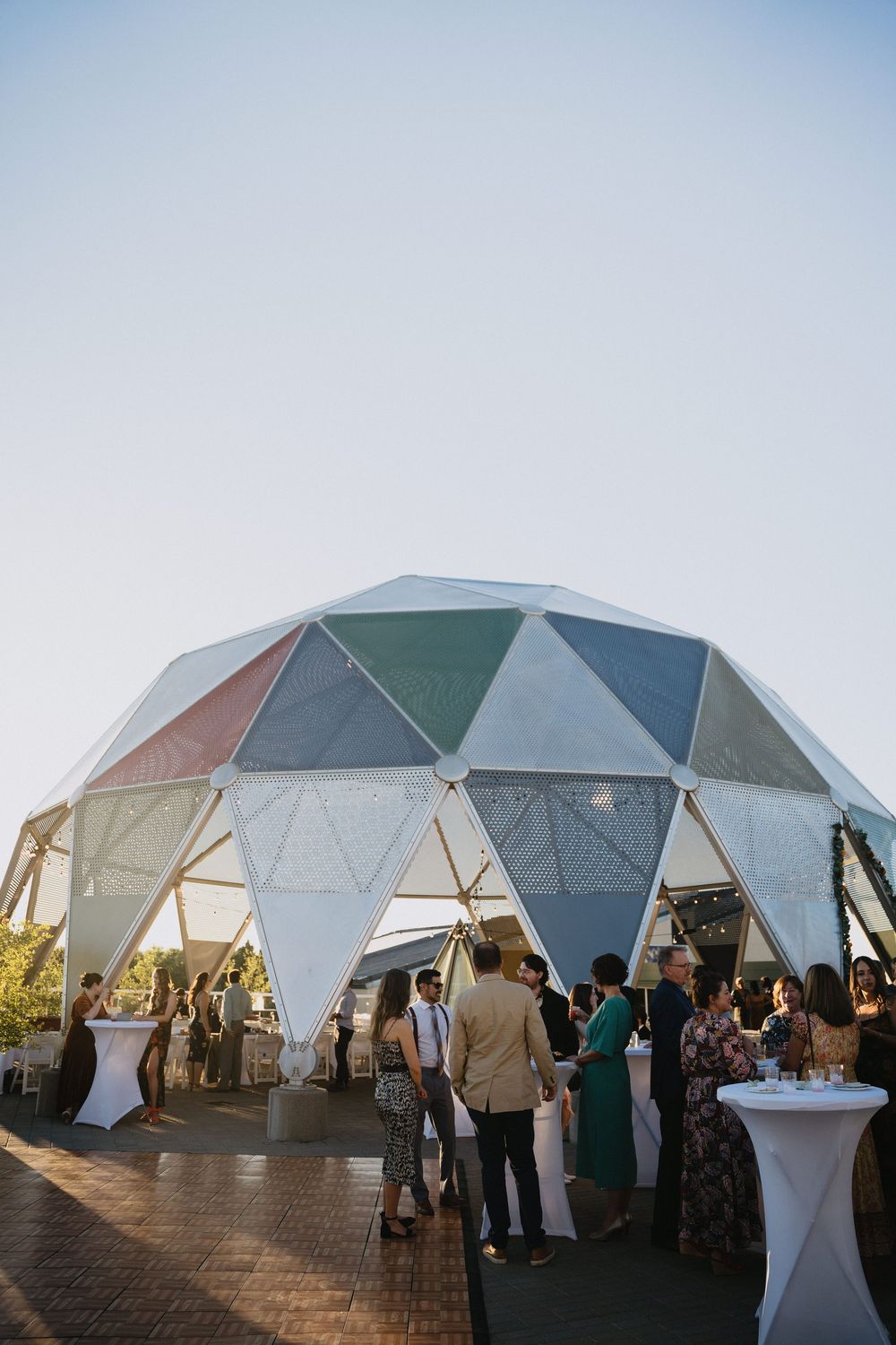 Colorful geodesic dome tent hosting an outdoor evening wedding reception with guests mingling on a brick patio.