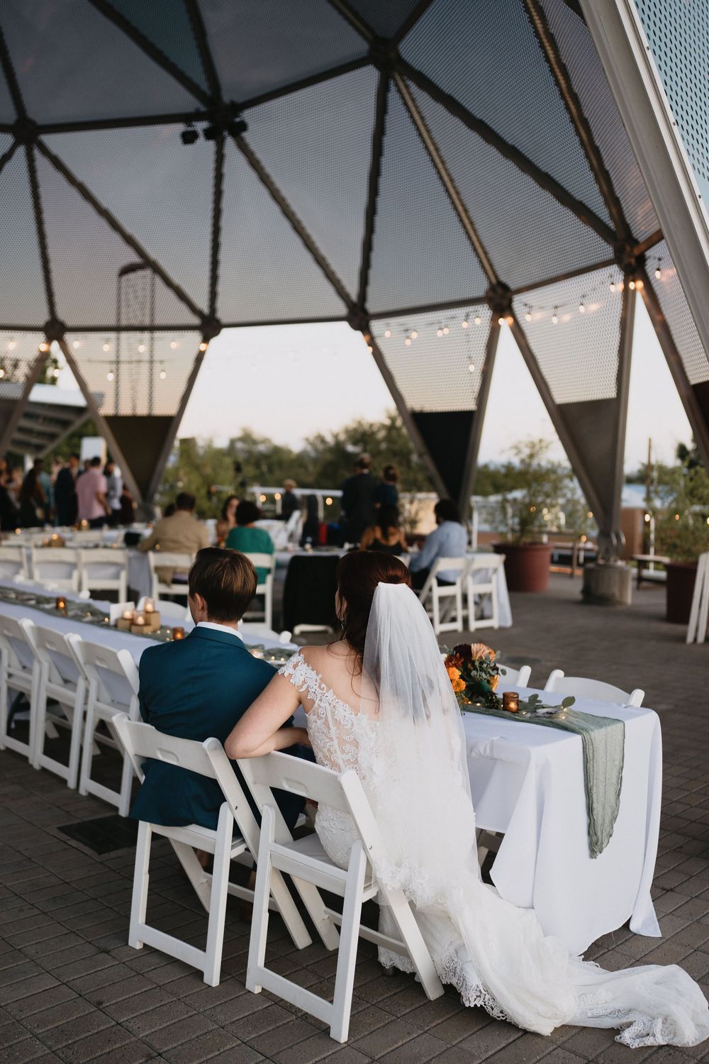 Bride and groom sit at elegant outdoor wedding reception table under geometric dome venue at dusk.