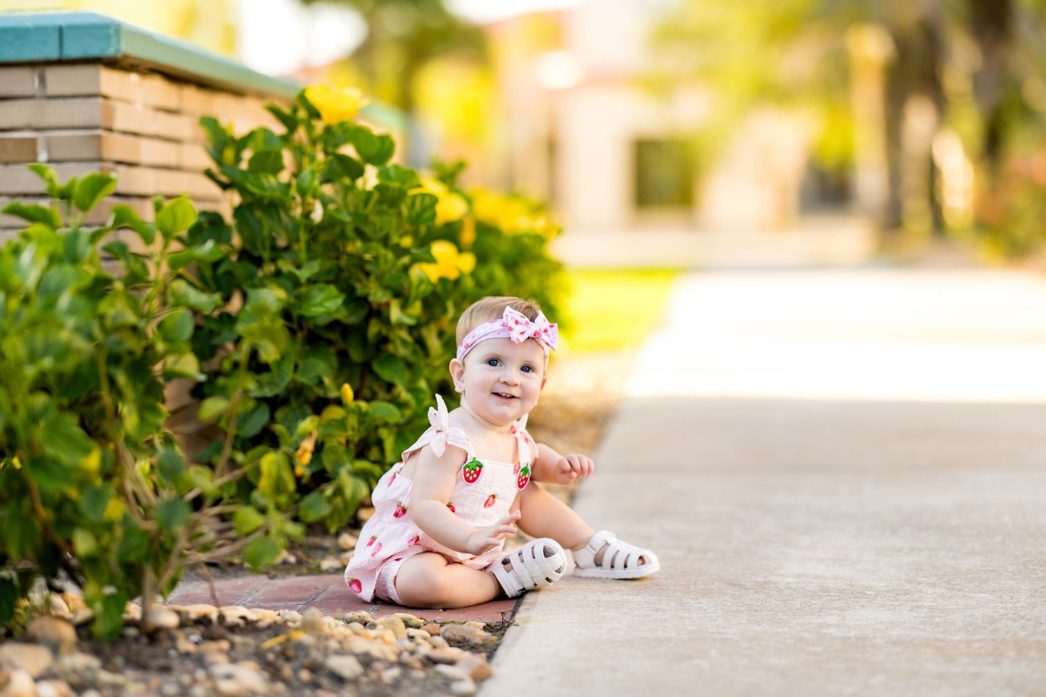 Adorable baby girl in floral romper and headband sitting on a sunny sidewalk next to green and yellow garden bushes.