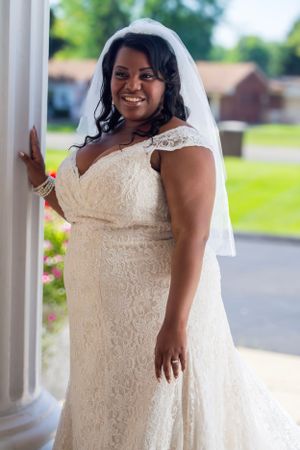 Bride in a lace cap sleeve wedding dress poses gracefully against a white column on her wedding day.