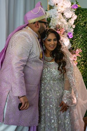 A couple in lavender and silver formal attire pose together at a floral decorated event backdrop.