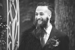 Joyful groom with full beard wearing dark suit and tie smiles during wedding celebration.