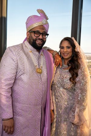 A couple in traditional South Asian wedding attire poses together, wearing lavender and silver embellished outfits.