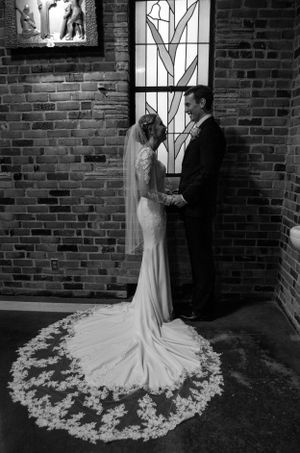 Elegant couple in wedding attire poses against brick wall with dramatic train on dress.