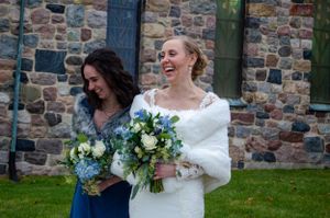 Two people in wedding attire holding bouquets laugh together outside stone building.