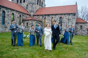Wedding party in blue attire posing outside historic stone church with red roof.