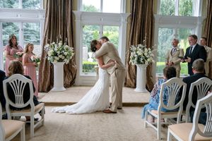 An intimate wedding ceremony takes place in a bright sunlit room with white chairs and floral arrangements by large windows.