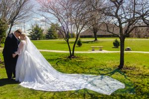 A wedding couple shares an intimate moment on a green lawn with a flowing white cathedral veil trailing behind them.