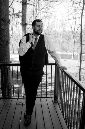 A formal portrait of a groom in a vest and tie standing on a wooden deck overlooking nature.