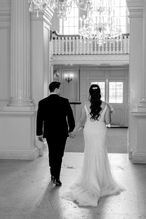A romantic black and white photo of newlyweds walking through an elegant hall with crystal chandeliers.
