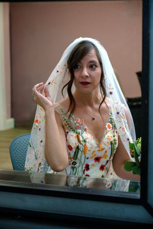 A bride wearing a floral embroidered wedding dress and veil poses behind a window.