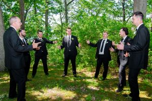 Groomsmen in black suits sharing a laugh in a wooded outdoor setting.