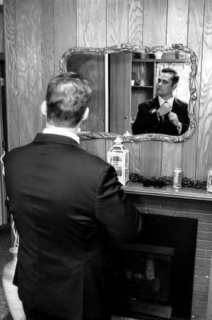 A man in a suit adjusts his tie while looking in an ornate vintage mirror.