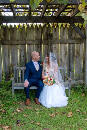 A couple in wedding attire sits together on a garden bench under bunting decorations and greenery.