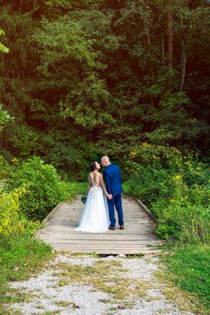 A couple in wedding attire walks along a wooden boardwalk path surrounded by lush green forest vegetation.